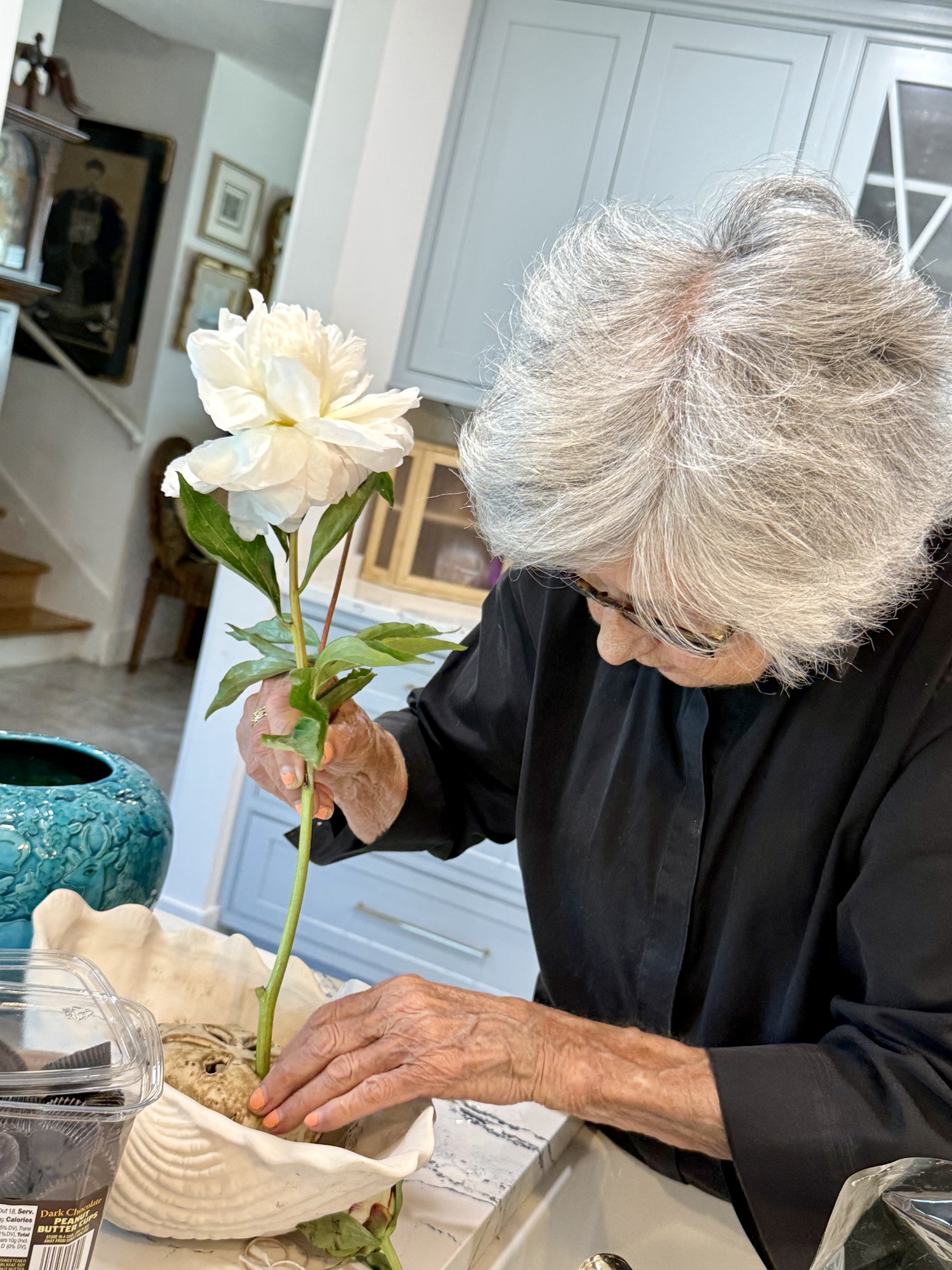 Close-up of an older woman's hands arranging a white peony in a shell-shaped bowl