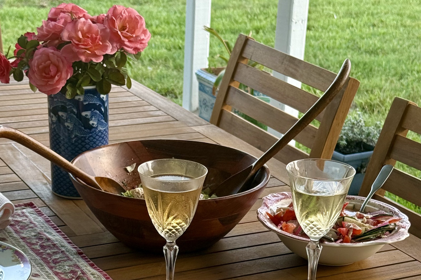 An outdoor table set with a wooden salad bowl, patterned dishes, wine glasses, and a vase of pink roses